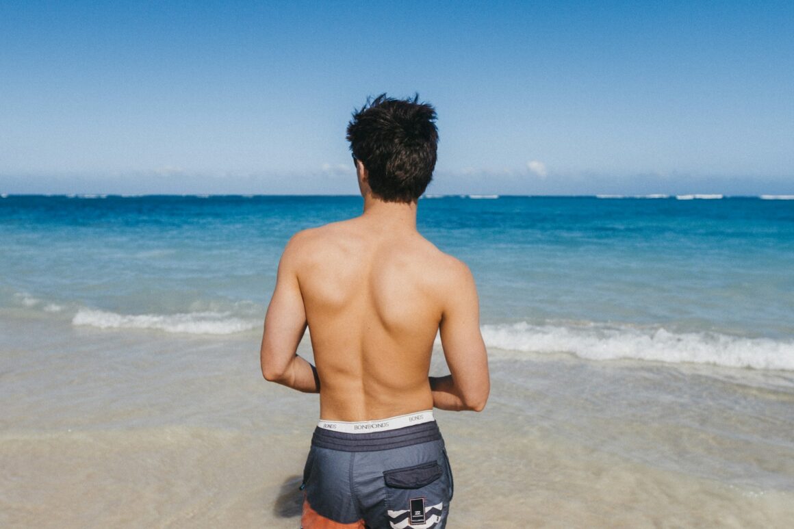 A guy standing on a sunny beach by the ocean on a hot day in swimwear