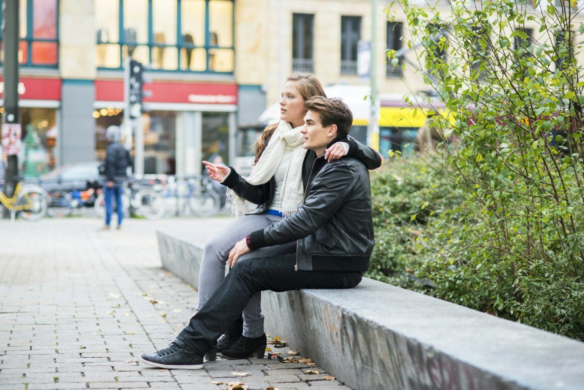 A teen guy at the height of puberty sitting on a low wall with his girlfriend on his lap