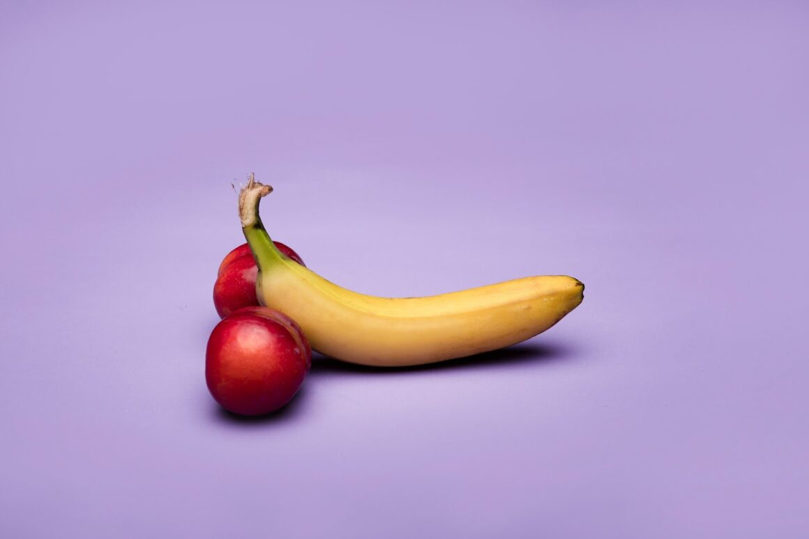 A banana with two plums to represent a penis and testicles on a purple aesthetic background