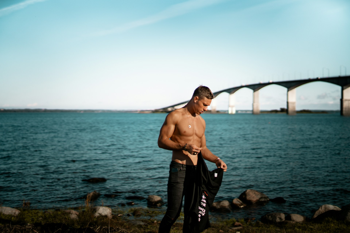 A shirtless ripped guy standing in front of a bridged river in an aesthetic manner