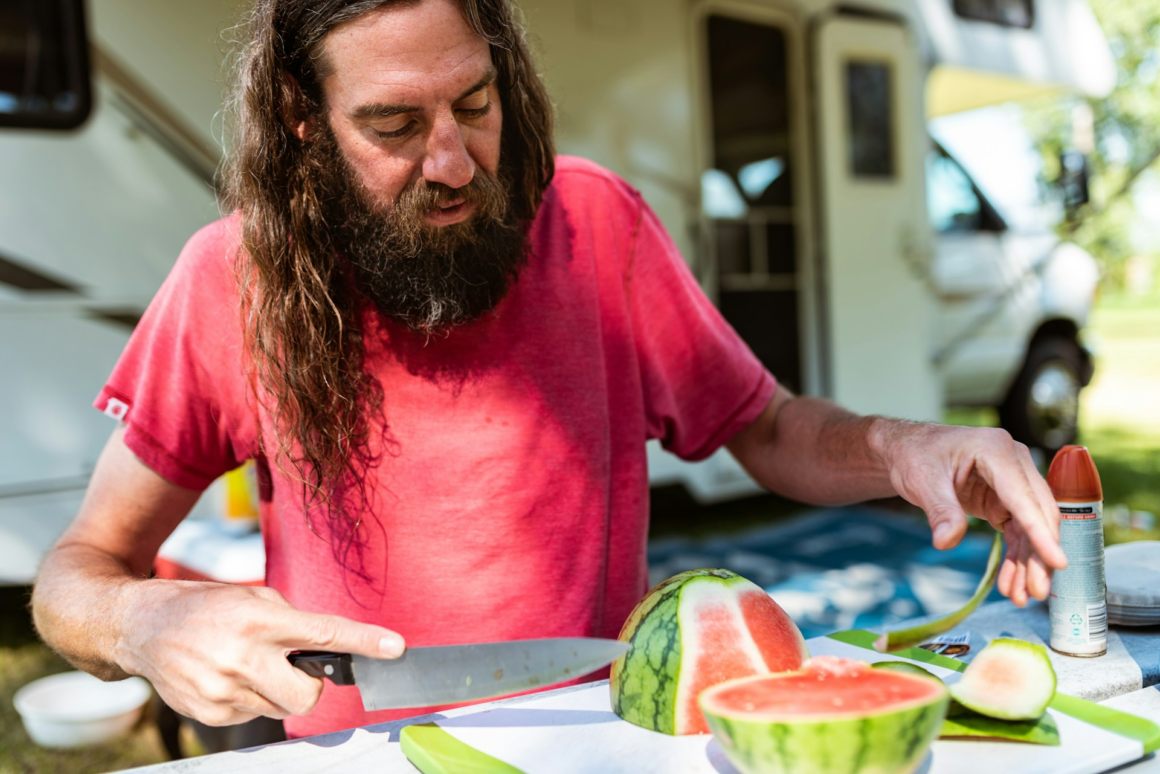 A man with long heair cutting and eating watermelon