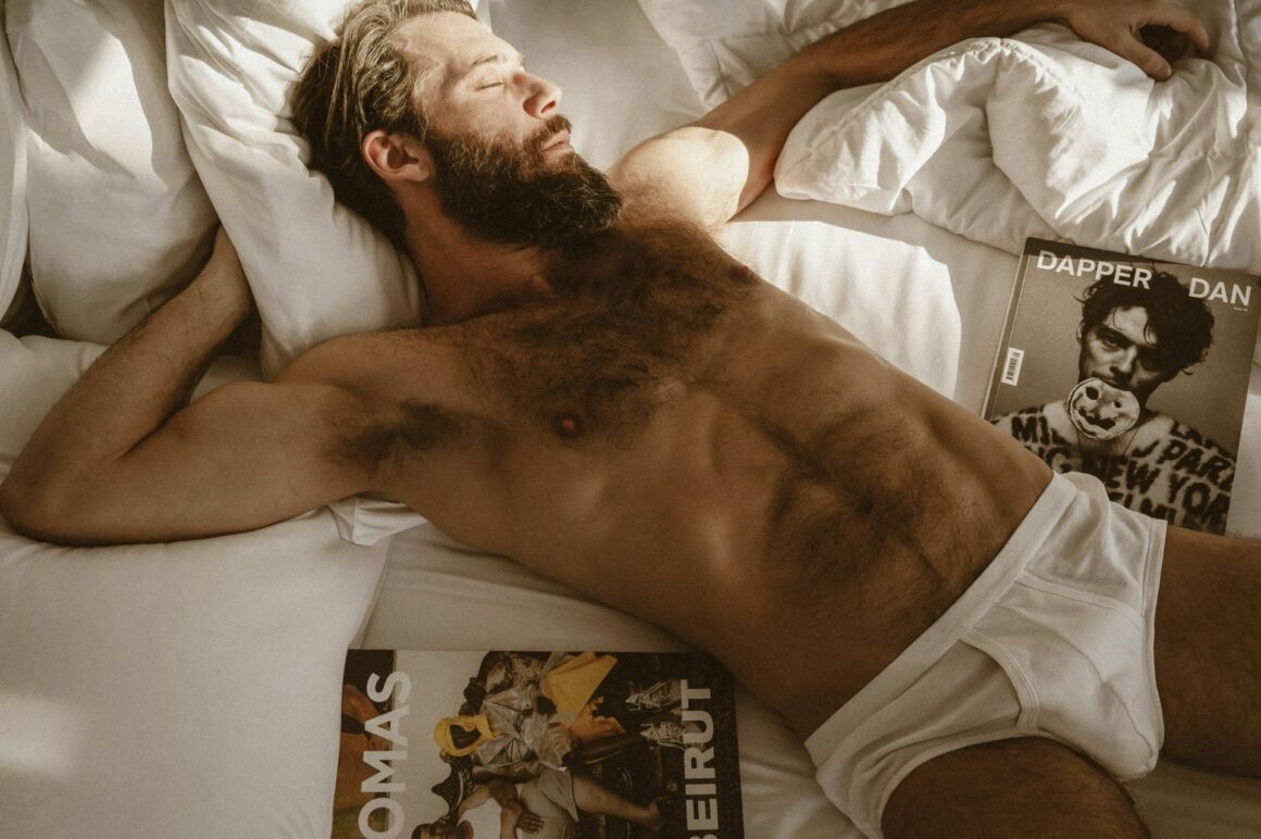 A shirtless man lying in bed with underwear on surrounded by magazines