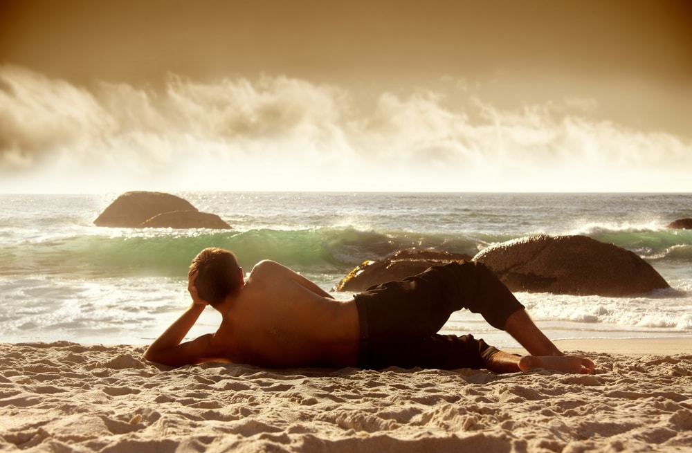A manscaped man laying on his side on a beach by a beautiful ocean.
