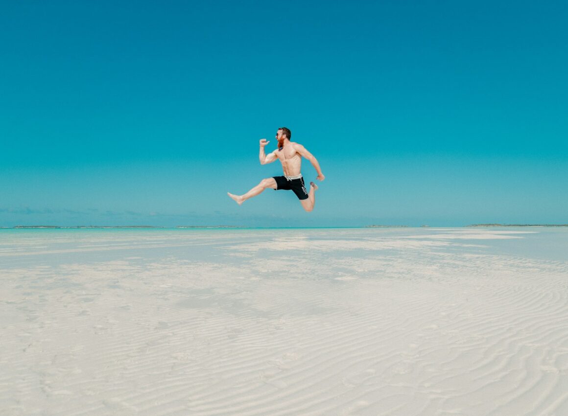 A man in shorts happy and jumping in the air at the beach on a bright blue day