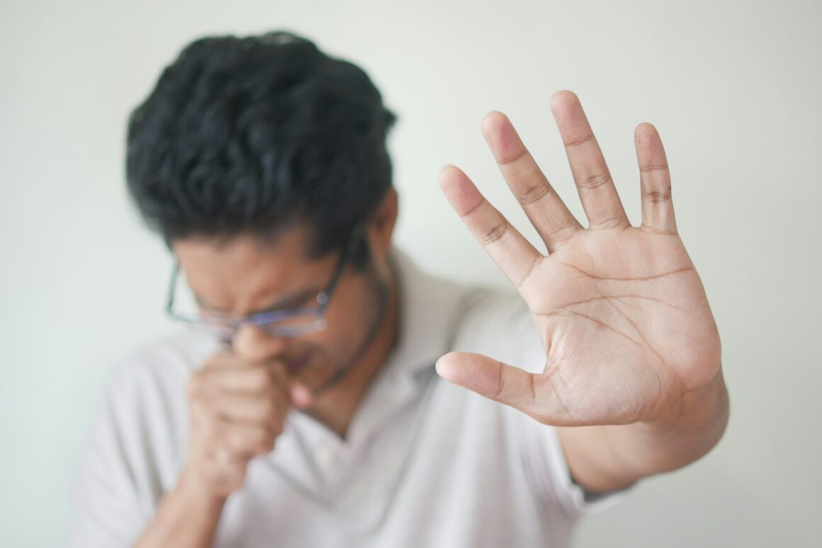 A man coughing and holding his palm up to the camera asking for no one to get close.