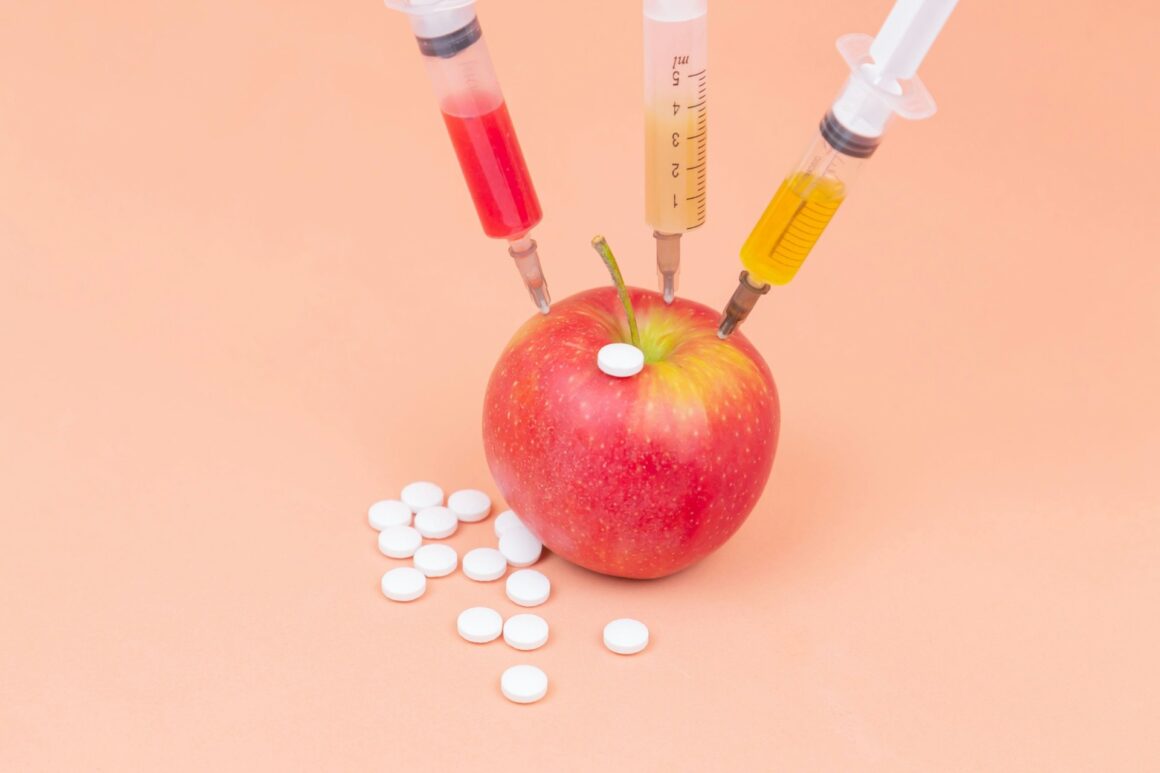 An apple with syringes stuck in it surrounded by little white pills