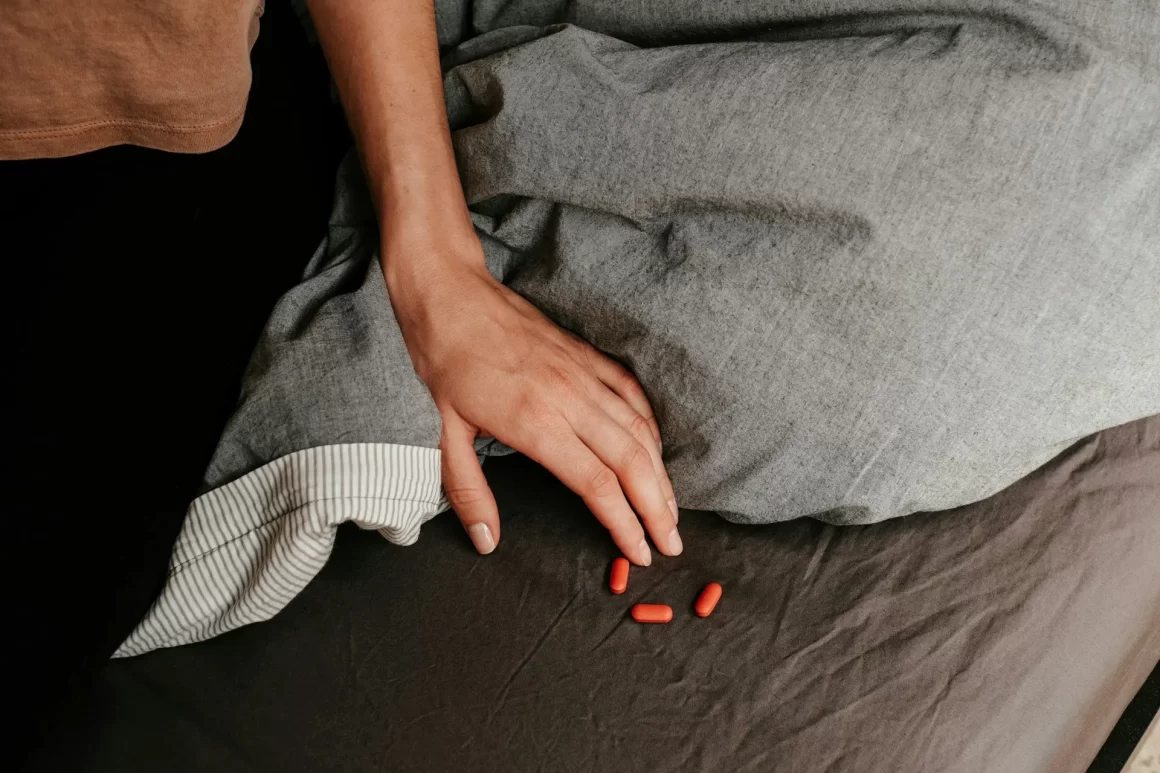 A woman reaching down to take three red pill capsules from the end of her bed.