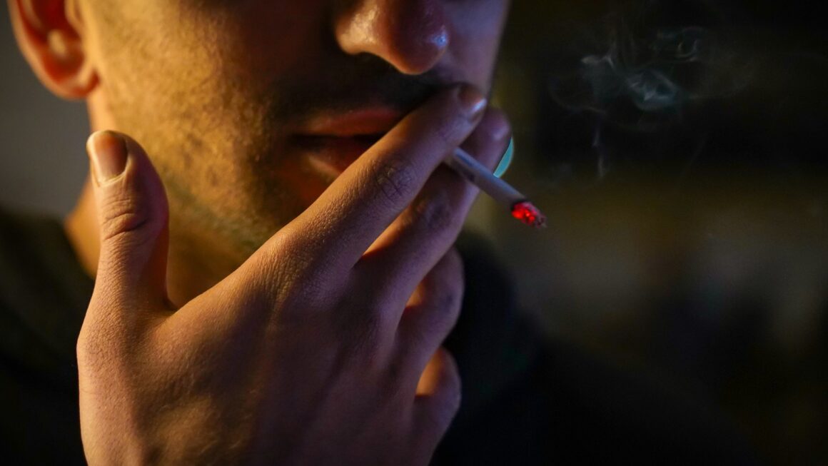 A close-up of a man smoking a cigarette outside and puffed tobacco smoke