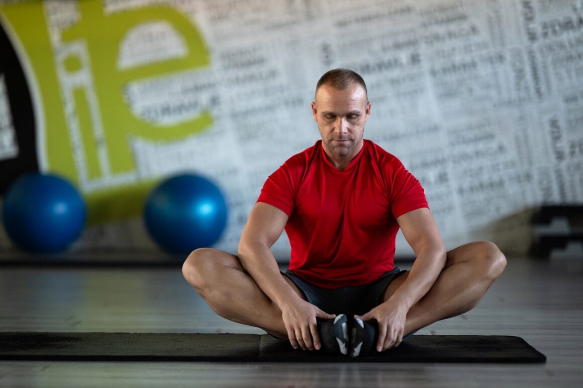 man wearing red shirt sitting on gym floor stretching